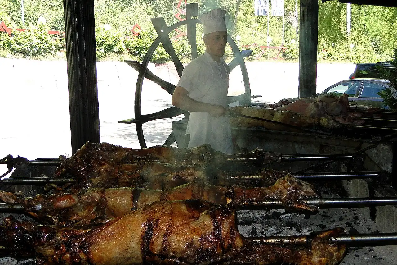 Whole lambs turning on spits over open coals at an outdoor grill, with a chef in a white uniform and toque tending the fire behind them