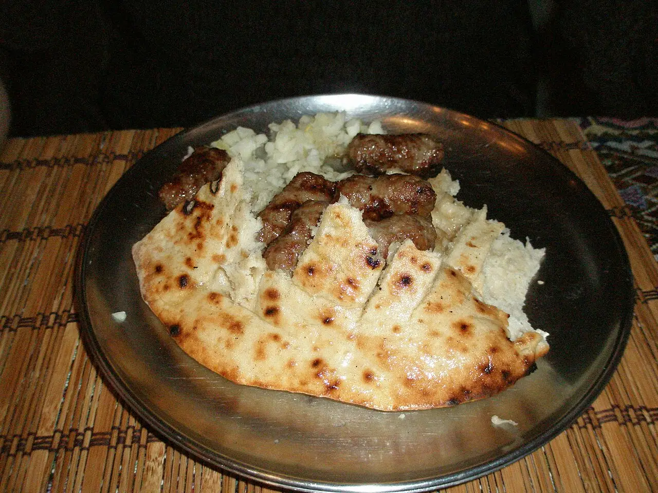 Grilled cevapi rolls in cut somun flatbread with a pile of chopped raw onion, served on a metal plate on a woven mat