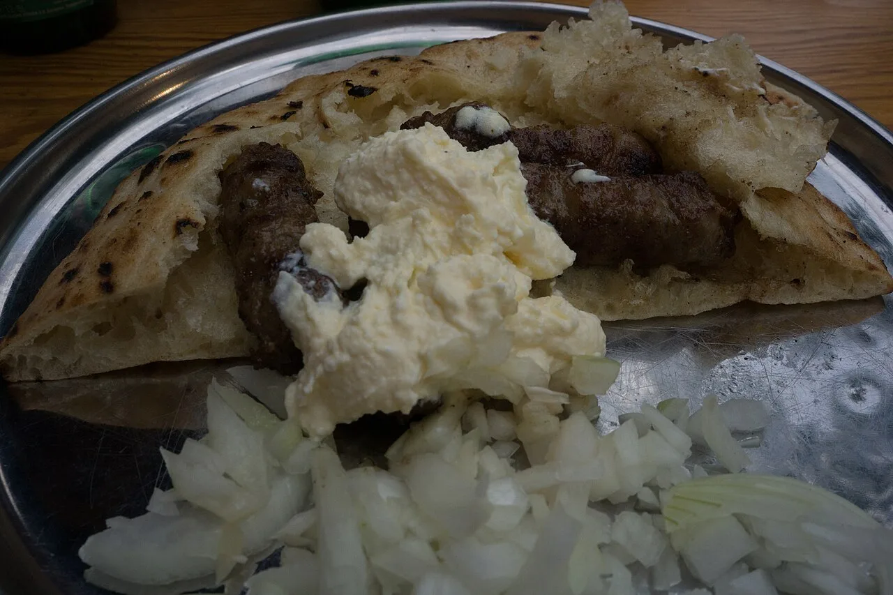 Grilled cevapi rolls tucked into somun flatbread with a scoop of white kajmak and a pile of chopped raw onion on a metal plate