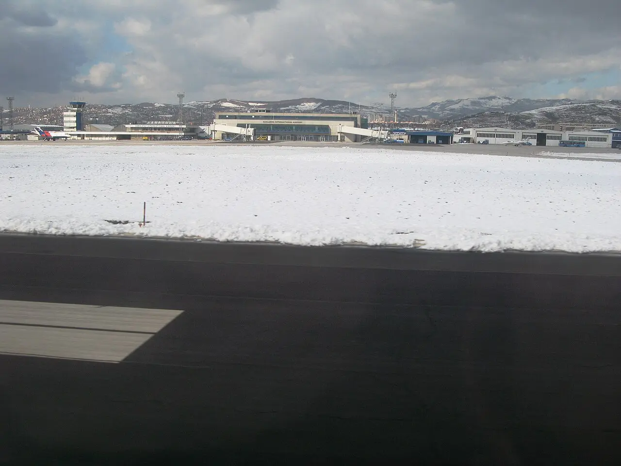 Sarajevo International Airport terminal in winter with snow on the apron and the mountains behind, showing how the airport sits in a valley bowl