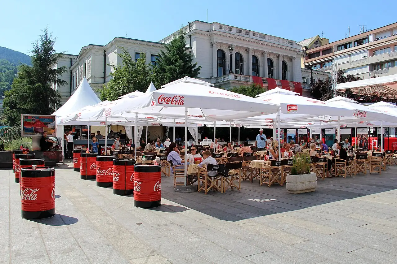 Festival Square cafe terraces with parasols in front of the grand cream facade of the National Theatre in Sarajevo on a sunny day