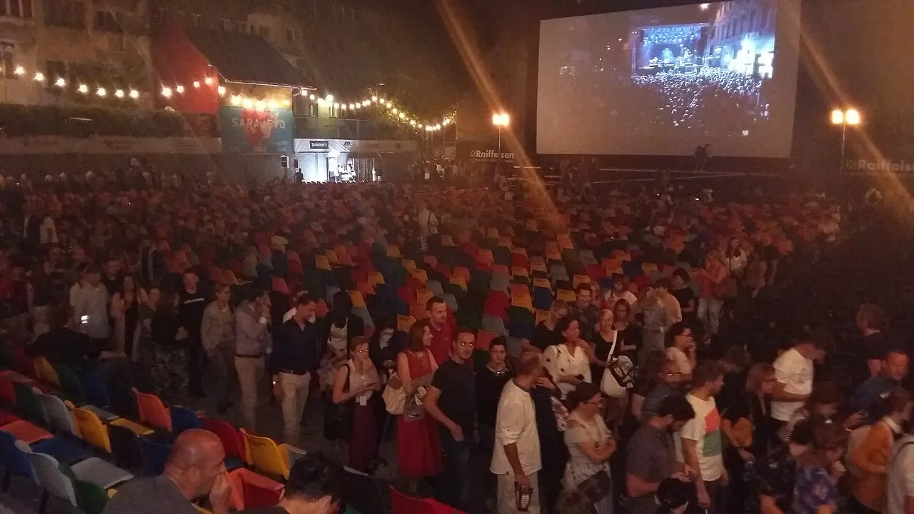 A packed night crowd in the Open Air Cinema at the Sarajevo Film Festival, colourful seats facing a huge lit screen under a Sarajevo heart sign