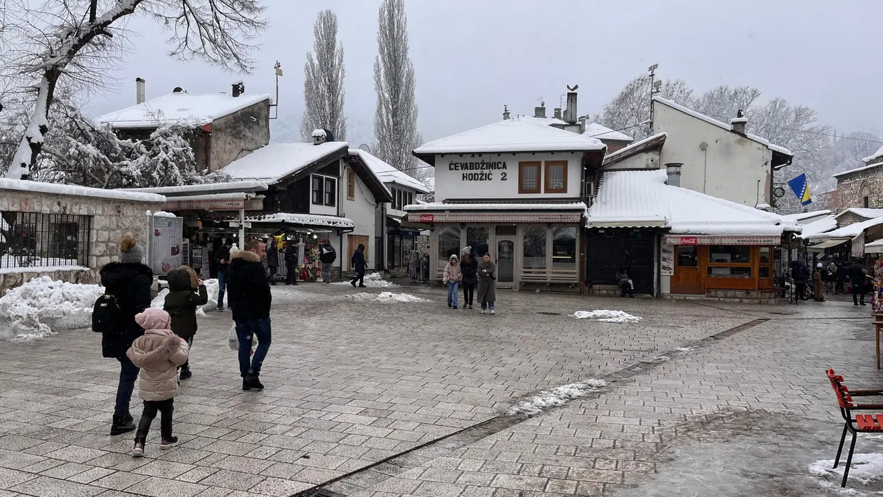 Snow on the roofs of the low wooden shops around a square in Baščaršija, Sarajevo, a family in winter coats crossing the wet paving and a snow-laden tree on the left