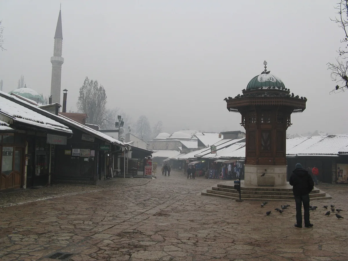 The wooden Sebilj fountain in Baščaršija on a grey winter morning, snow on the shop roofs and the minaret behind fading into haze, pigeons on the paving