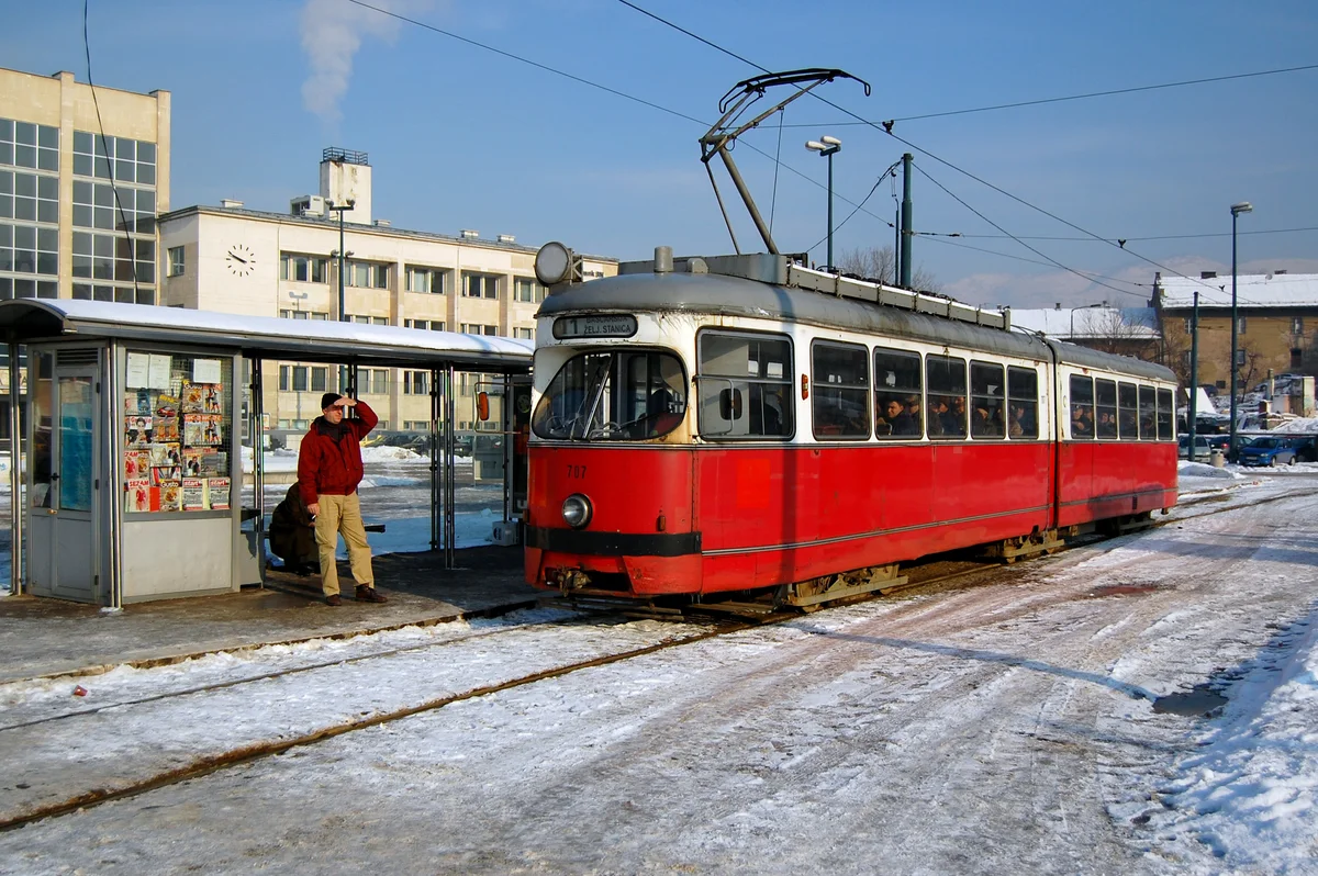 A red and white Sarajevo tram signed Line 1 to the railway station at a snowy stop on a clear day, a man in a red jacket by the newspaper kiosk on the platform