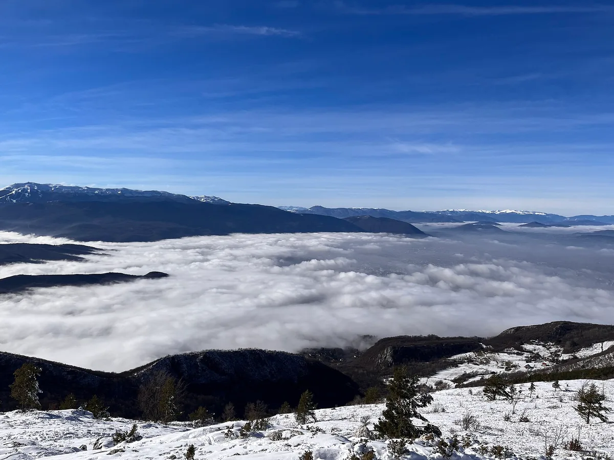 View from snowy Trebević under a deep blue sky, the Sarajevo valley below completely hidden by a flat sea of white fog, snow-capped ranges on the horizon