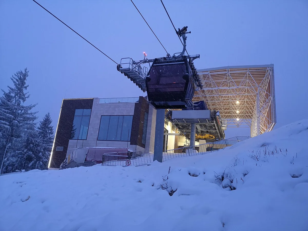 A gondola of the Trebević cable car arriving at a lit station at dusk in falling snow, snow-covered spruces on the left