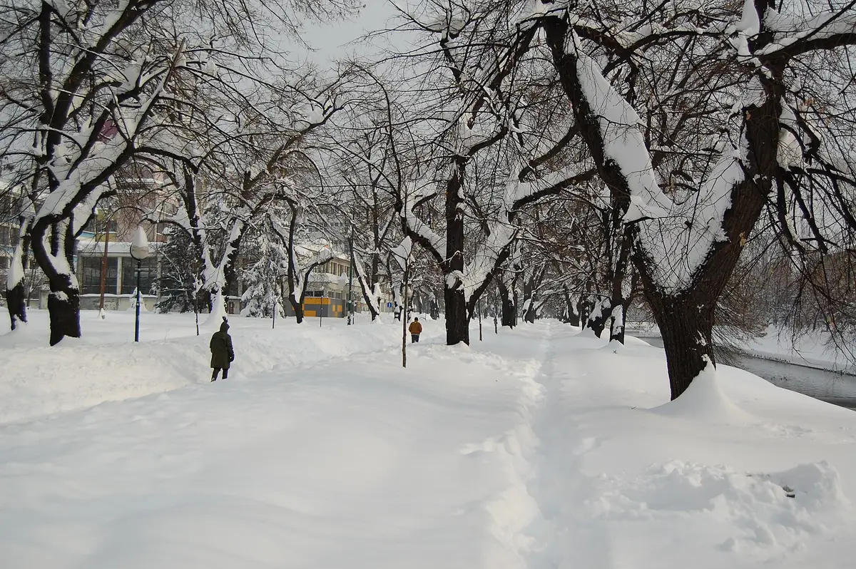 Wilson's Promenade in Sarajevo under deep snow, two walkers on a trodden path between rows of snow-laden trees, the Miljacka river on the right