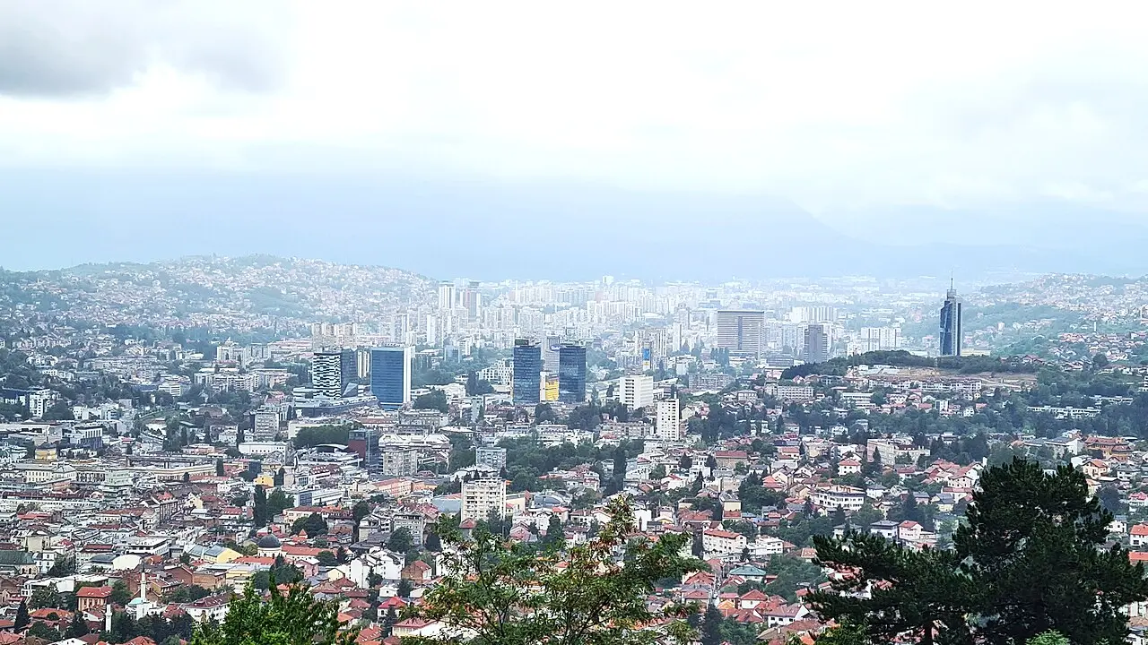 Sarajevo spread across its narrow valley at dusk, lights coming on between minarets and tower blocks under wooded ridges