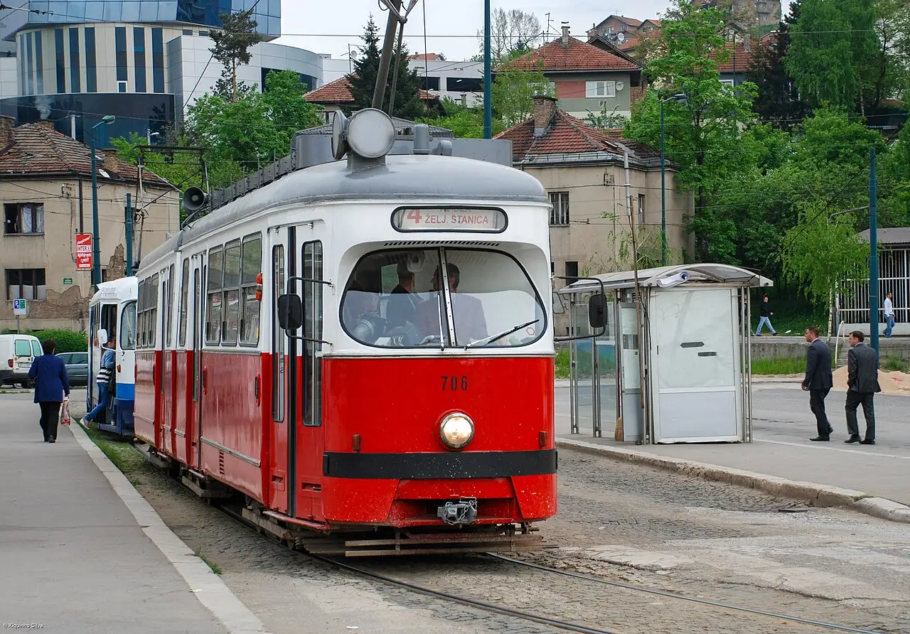 An older red Sarajevo tram on the valley tracks with passengers nearby