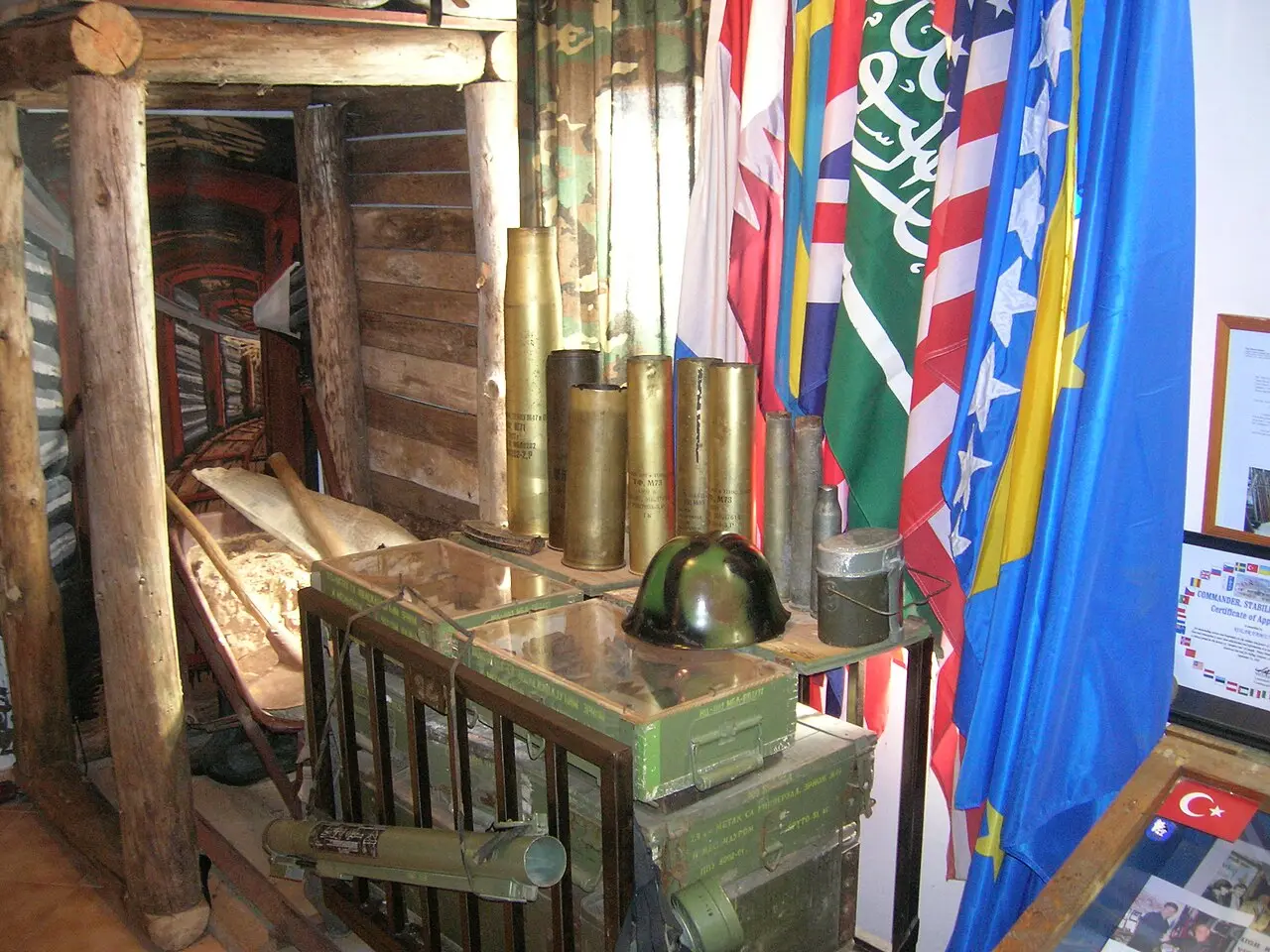 The low, wood-lined Sarajevo tunnel passage receding into the distance, with wartime exhibits and donor-country flags at the entrance