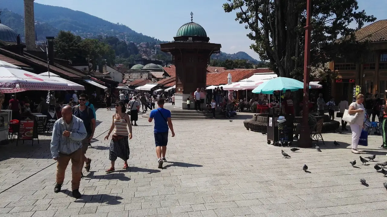 The wooden Sebilj fountain on Baščaršija square in Sarajevo under a bright sky, pigeons and café parasols around the cobbles