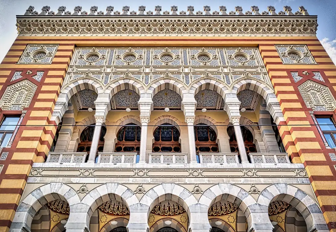 The striped pseudo-Moorish facade of Sarajevo City Hall (Vijećnica) beside the Miljacka under a clear sky