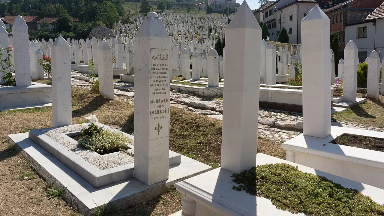 Rows of white gravestones on a green Sarajevo hillside at the Kovači memorial cemetery, the city below