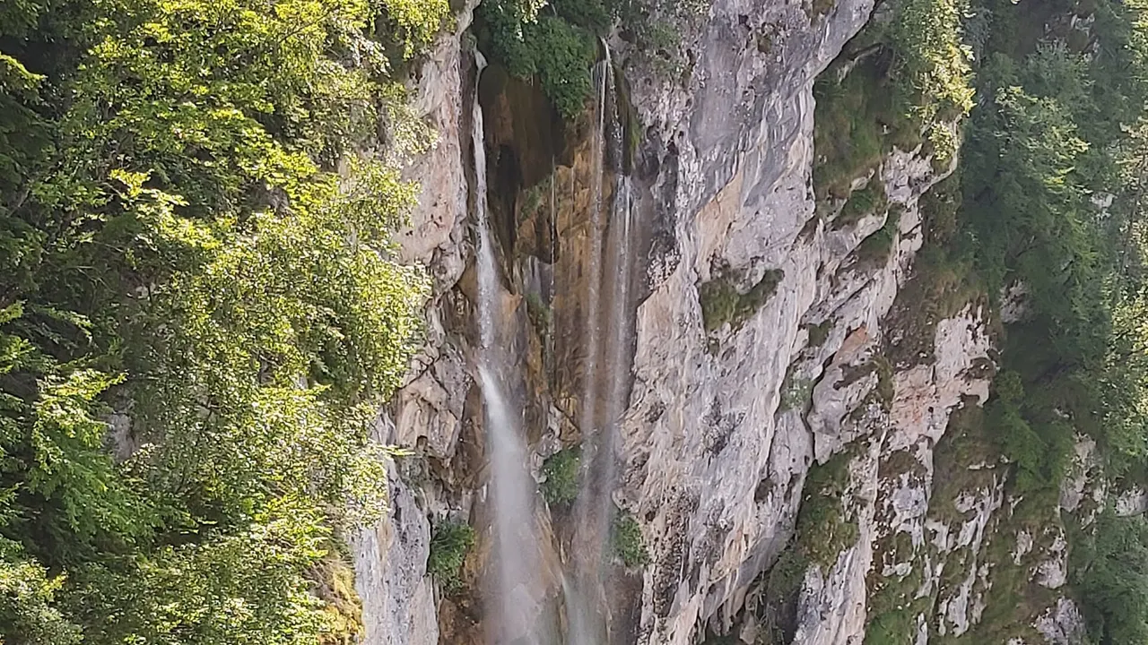 Skakavac waterfall north of Sarajevo, a single thread of water dropping down an ochre-streaked limestone cliff with sunlit beech woods on the left