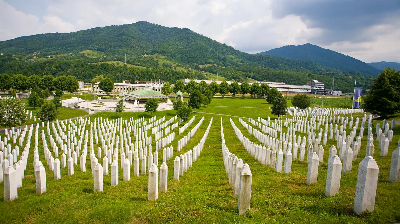The Srebrenica-Potočari cemetery: rows of white marble grave markers on green ground with the memorial pavilion and wooded hills behind