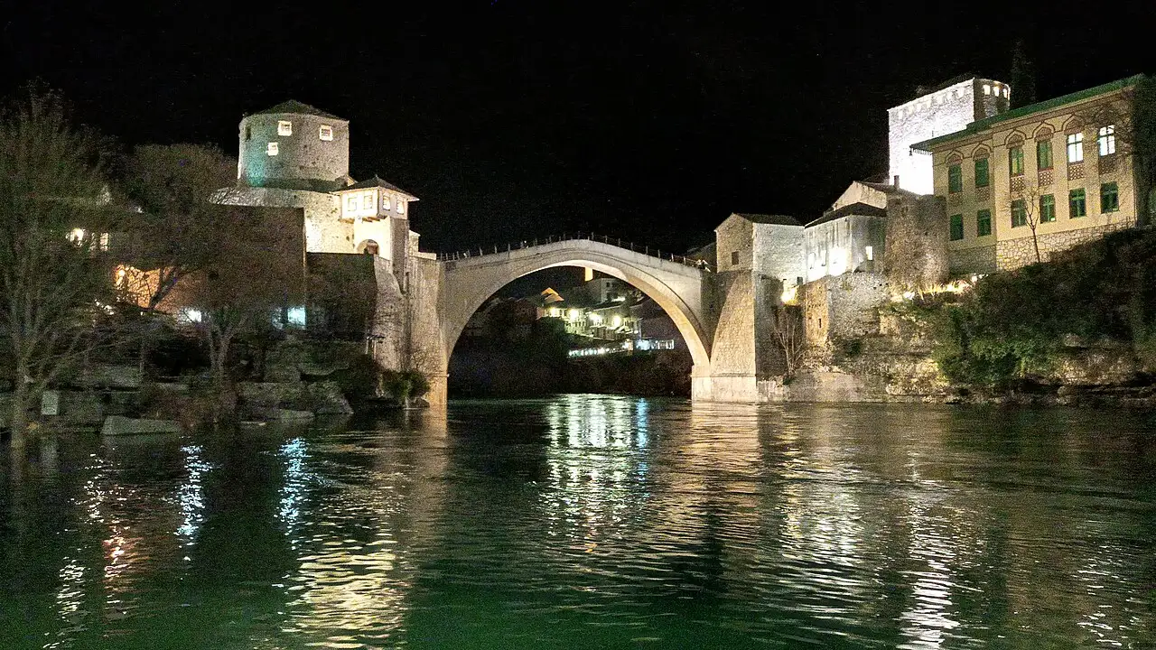 Stari Most lit up at night, its arch and the two flanking towers reflected in the dark Neretva