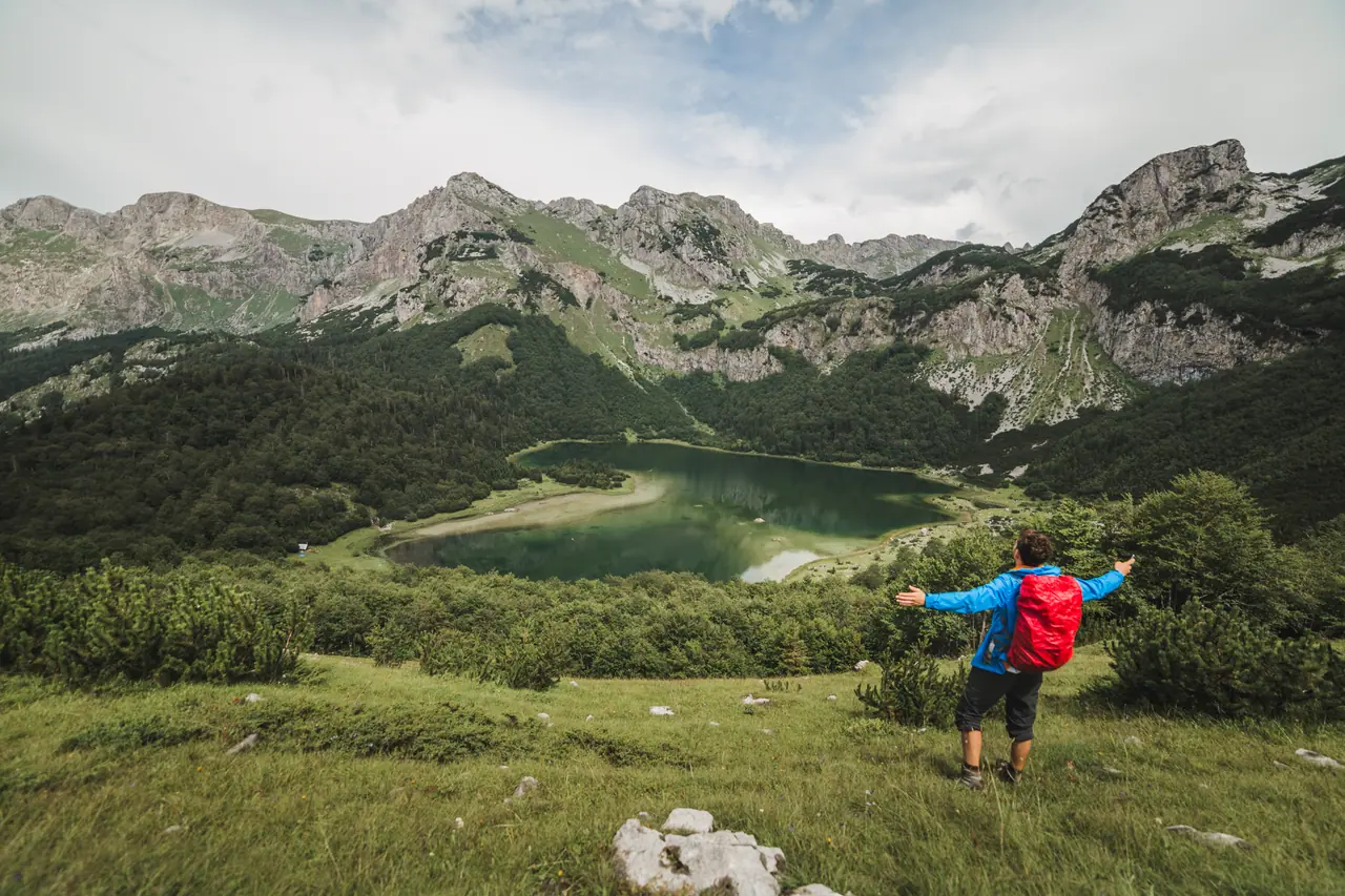 A hiker looking over Trnovačko lake, a green heart-shaped glacial lake ringed by the grey ridges below Maglić