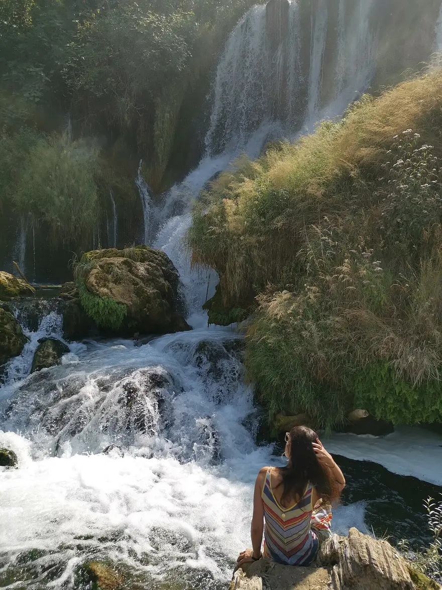A visitor sitting on a rock watching the Kravice waterfalls tumble over mossy tufa into a green pool