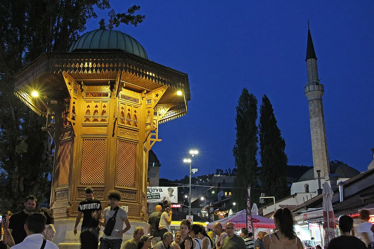 The illuminated wooden Sebilj fountain in Sarajevo's Baščaršija at dusk, crowds around it and a lit minaret behind