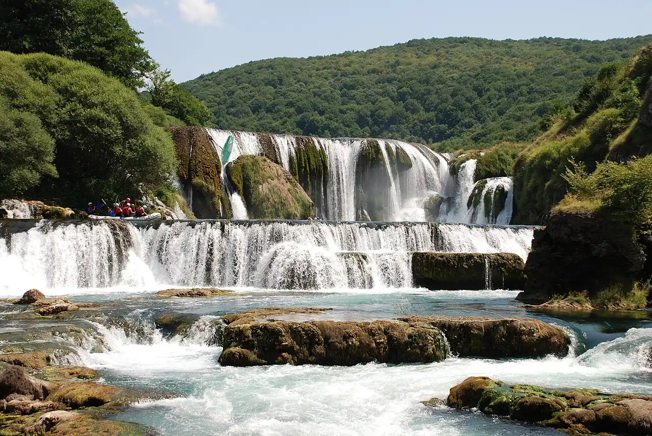 A raft of paddlers pausing at the lip of the wide terraced Štrbački buk waterfall on the turquoise Una river, forest all around