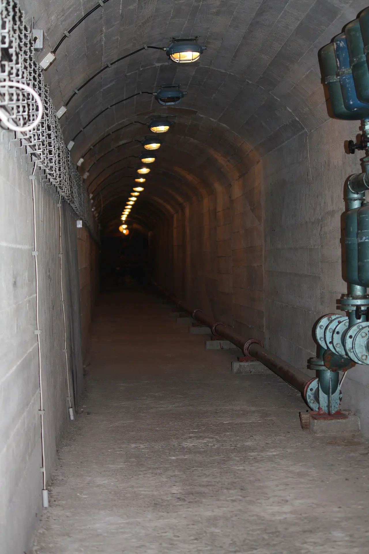 A long concrete access tunnel into the ARK D-0 bunker lined with pipework and dim wall lights