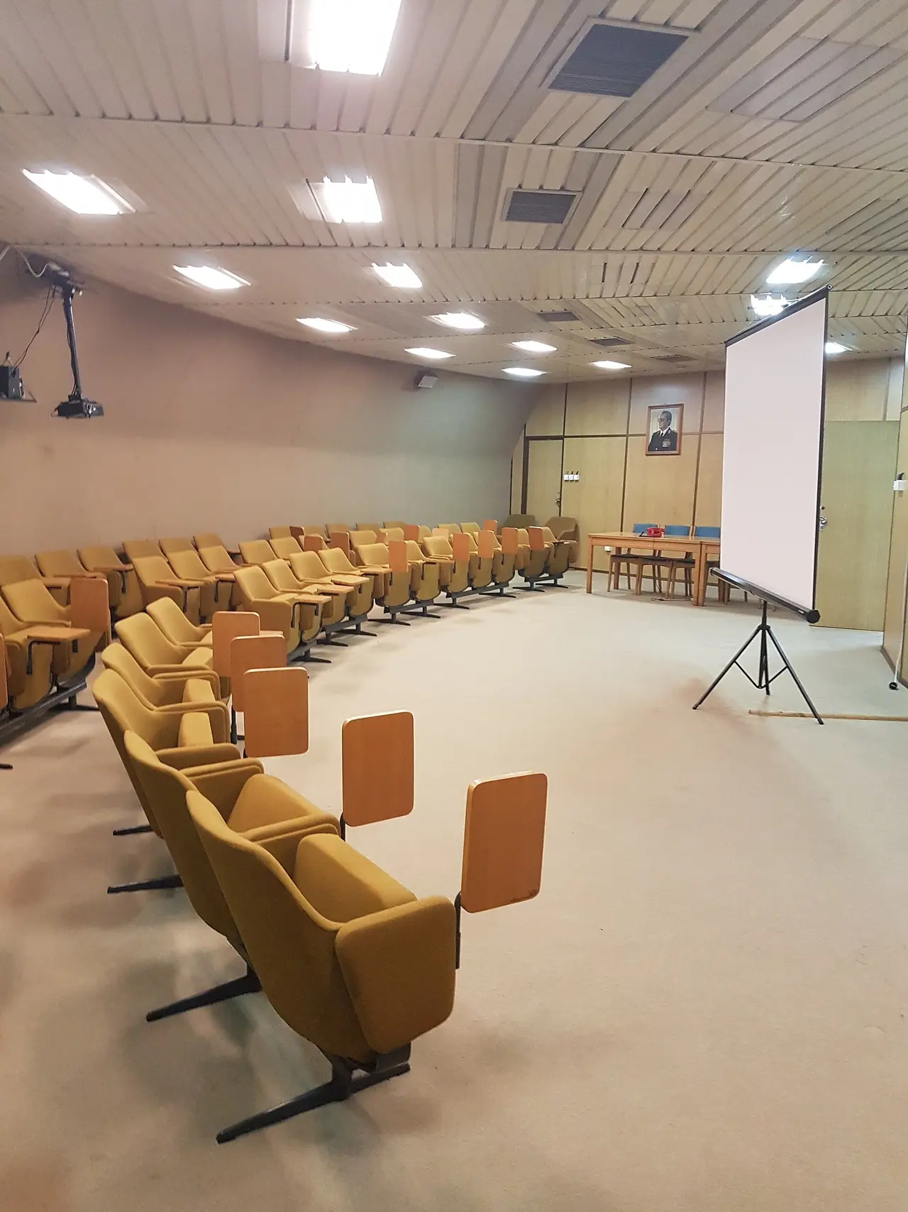 A briefing room inside the bunker with rows of yellow seats, a projector screen and a portrait of Tito on the wall