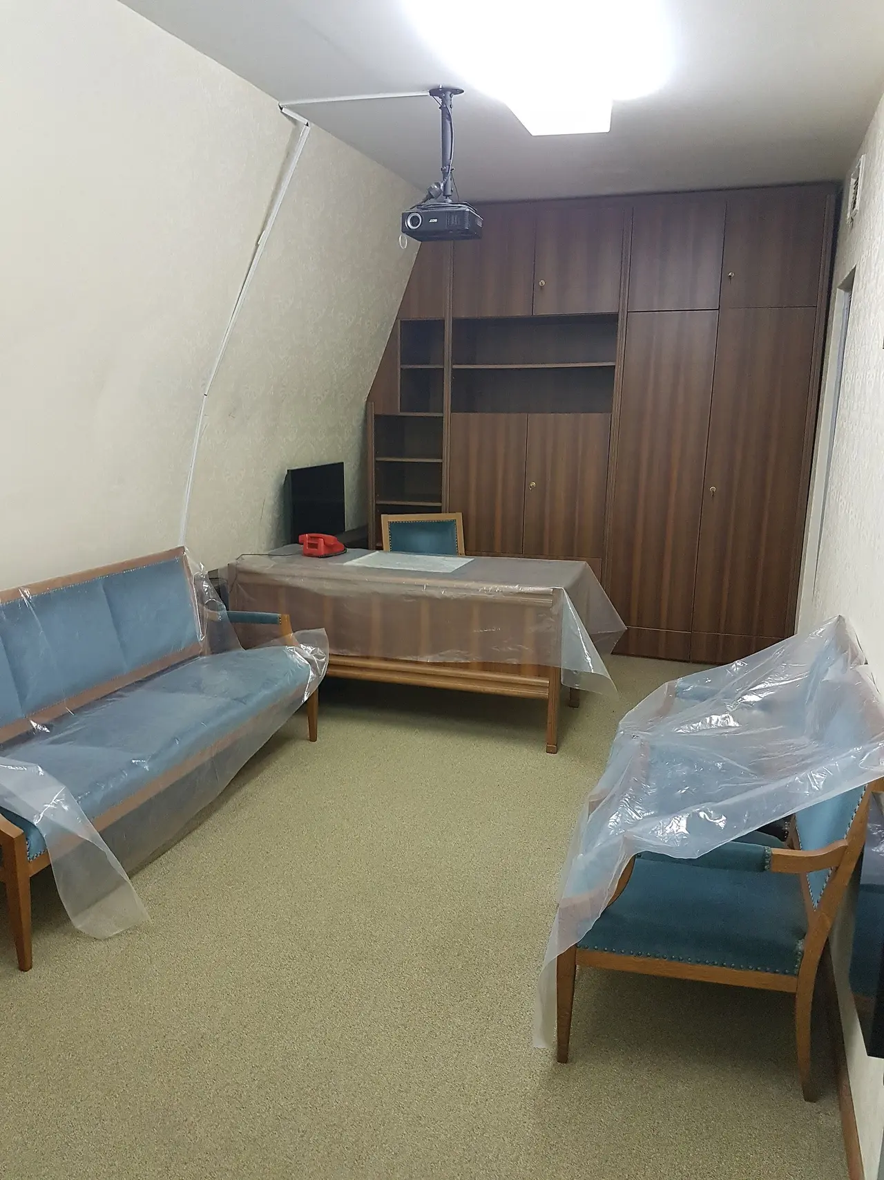 A residential room in the bunker with blue-upholstered furniture under plastic dust sheets and dark wood cabinets