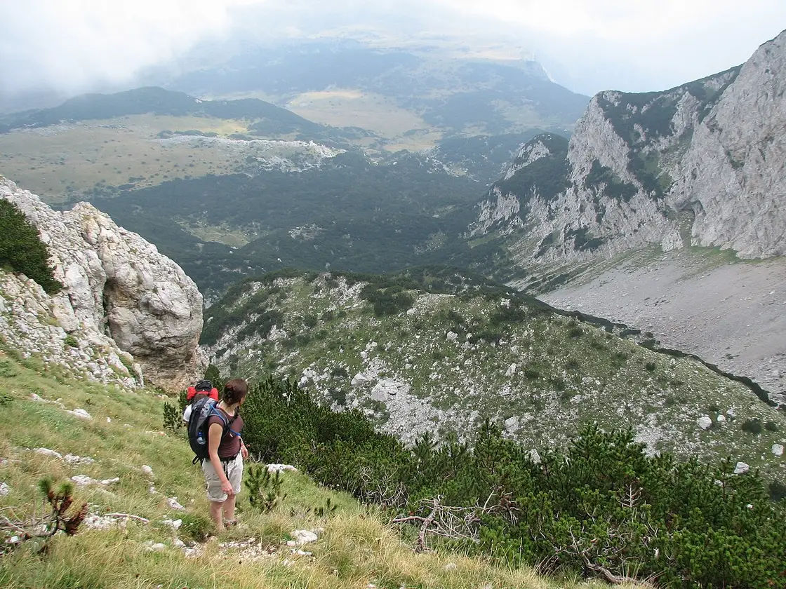 Hikers climbing a rocky ridge on Maglić with the deep green Sutjeska valley and forest far below