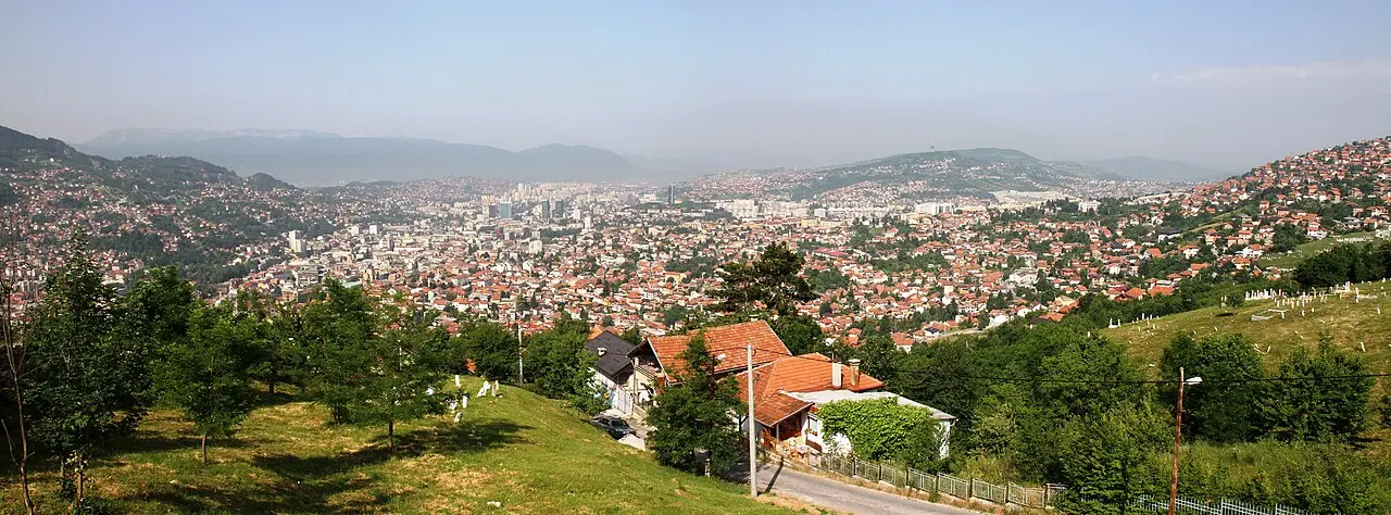 A wide panorama of Sarajevo filling the valley, minarets and apartment blocks with green hills rising on both sides