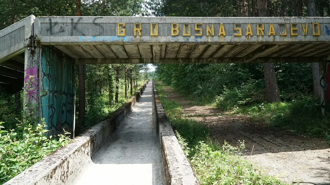 A graffitied concrete gantry over the straight run of the abandoned Sarajevo bobsled track disappearing into the pines