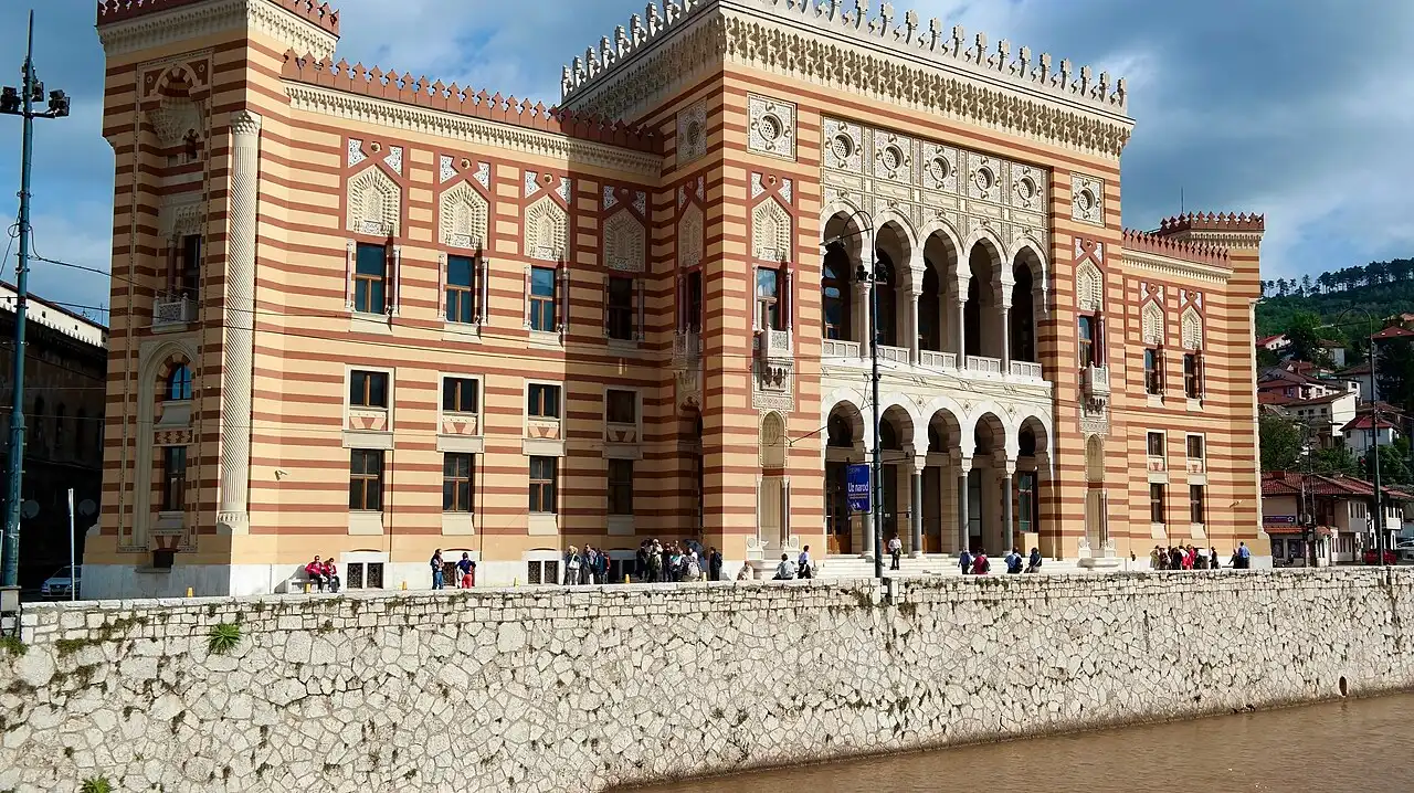The striped pseudo-Moorish facade of Sarajevo City Hall (Vijećnica) beside the Miljacka river under a clear sky