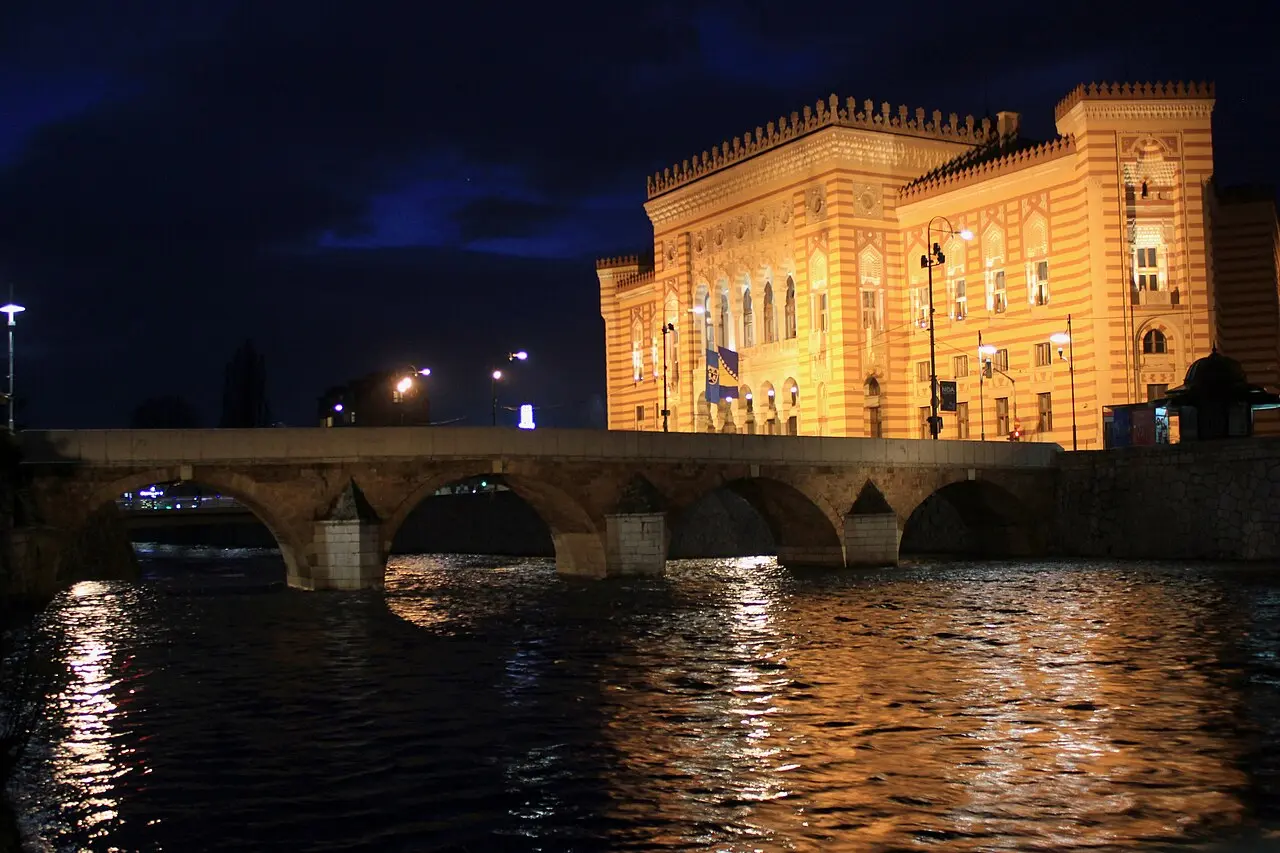 Vijećnica lit in warm floodlights at night, reflected in the Miljacka
