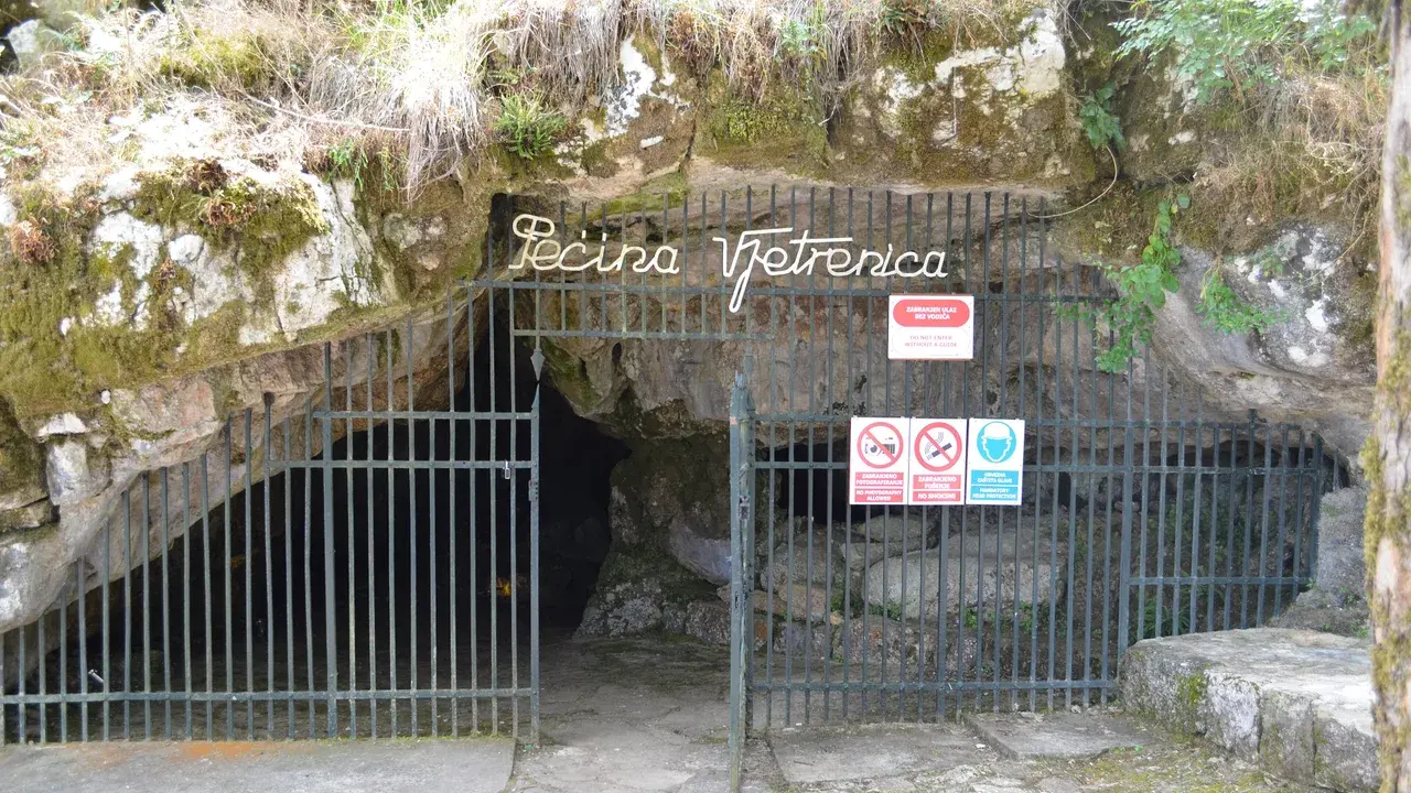 The gated entrance to Vjetrenica cave under a mossy limestone overhang, with the words Pecina Vjetrenica in metal letters above the iron gate