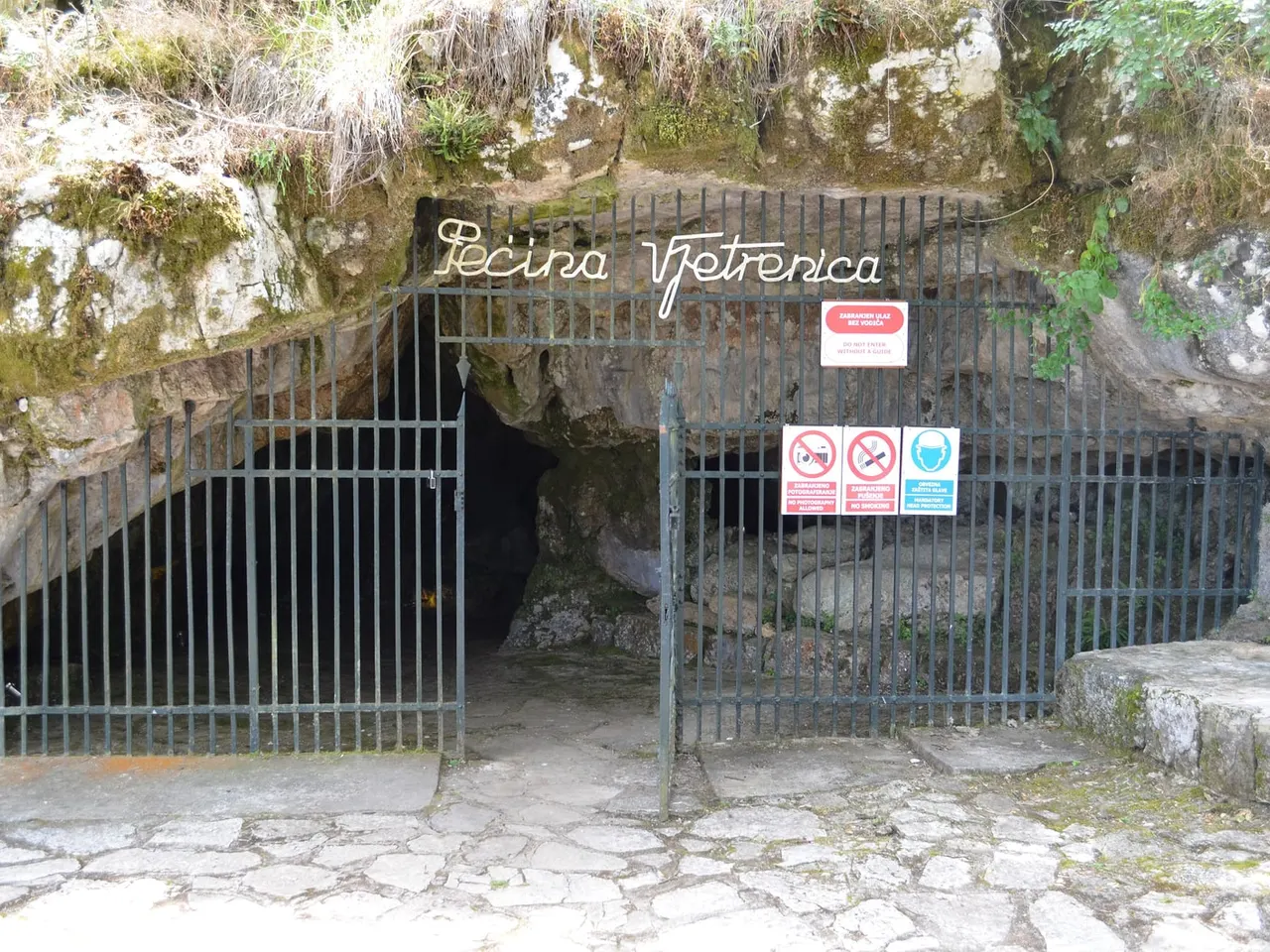 The locked iron gate across the mouth of Vjetrenica, the words Pecina Vjetrenica in metal letters above it and a red no-entry sign and helmet pictogram fixed to the bars