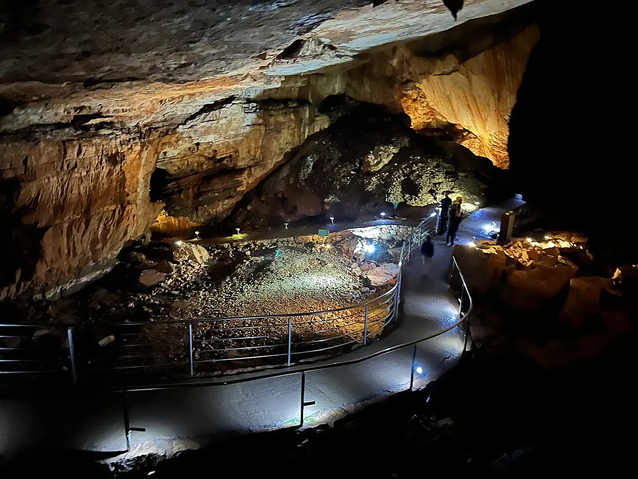 The concrete visitor path in Vjetrenica curving around a rockfall, lit by ground lamps under a broad limestone ceiling