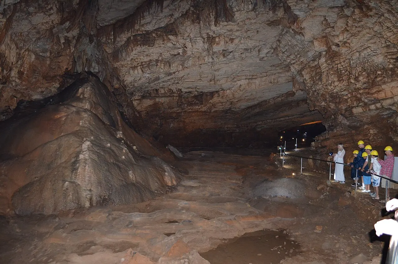 A wide low hall inside Vjetrenica with a group of visitors in yellow helmets standing at a metal railing beside the lit path