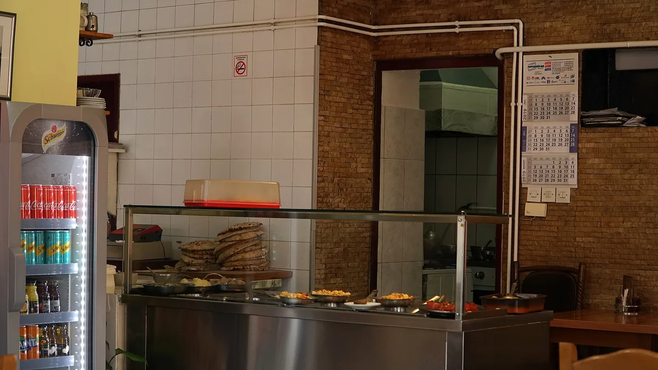 Inside a traditional Sarajevo aščinica: a stainless-steel counter with trays of stewed dishes and a stack of somun flatbreads under glass