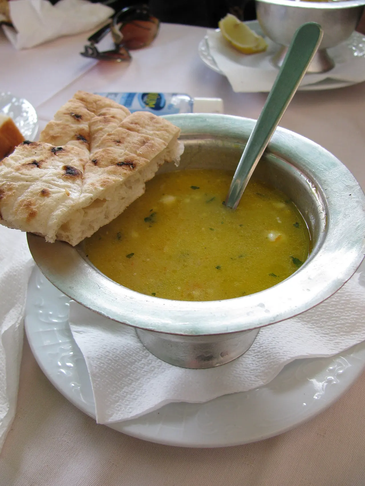 A metal bowl of begova čorba, a thick yellow Bosnian chicken and okra soup, served with a piece of somun flatbread