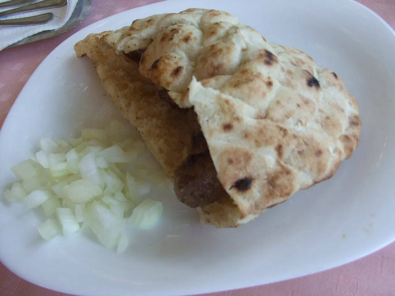 A portion of ćevapi in a folded somun flatbread with a pile of chopped raw onion on a plate in Mostar