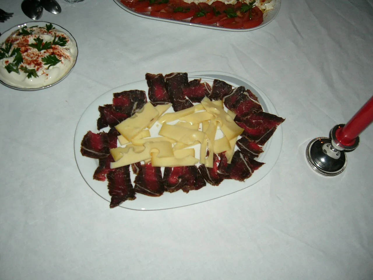A platter of thinly sliced Bosnian suho meso dry-cured beef arranged around slices of cheese, with kajmak on the side