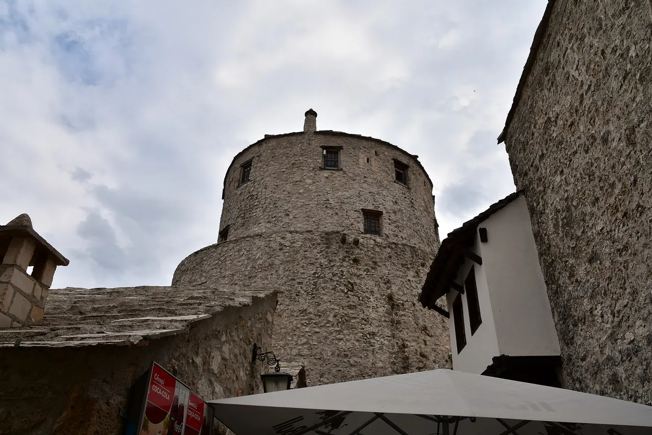 A red Coca-Cola sign reading Cevapi on a stone wall under Tara tower, with a restaurant umbrella in the foreground