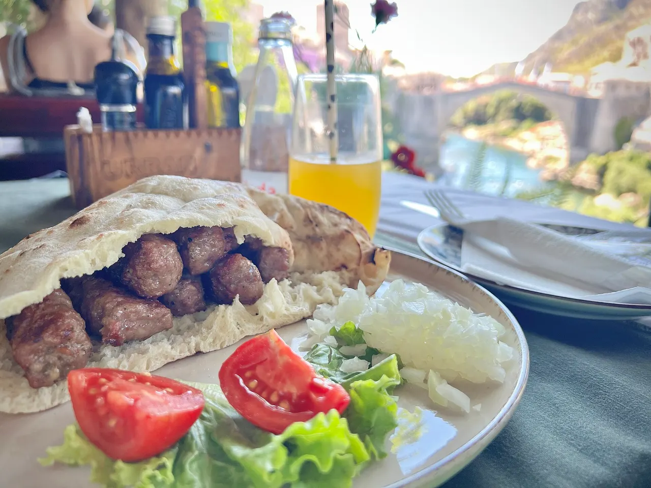 A plate of grilled cevapi in somun with chopped onion and tomato, Stari Most visible out of focus behind the terrace table