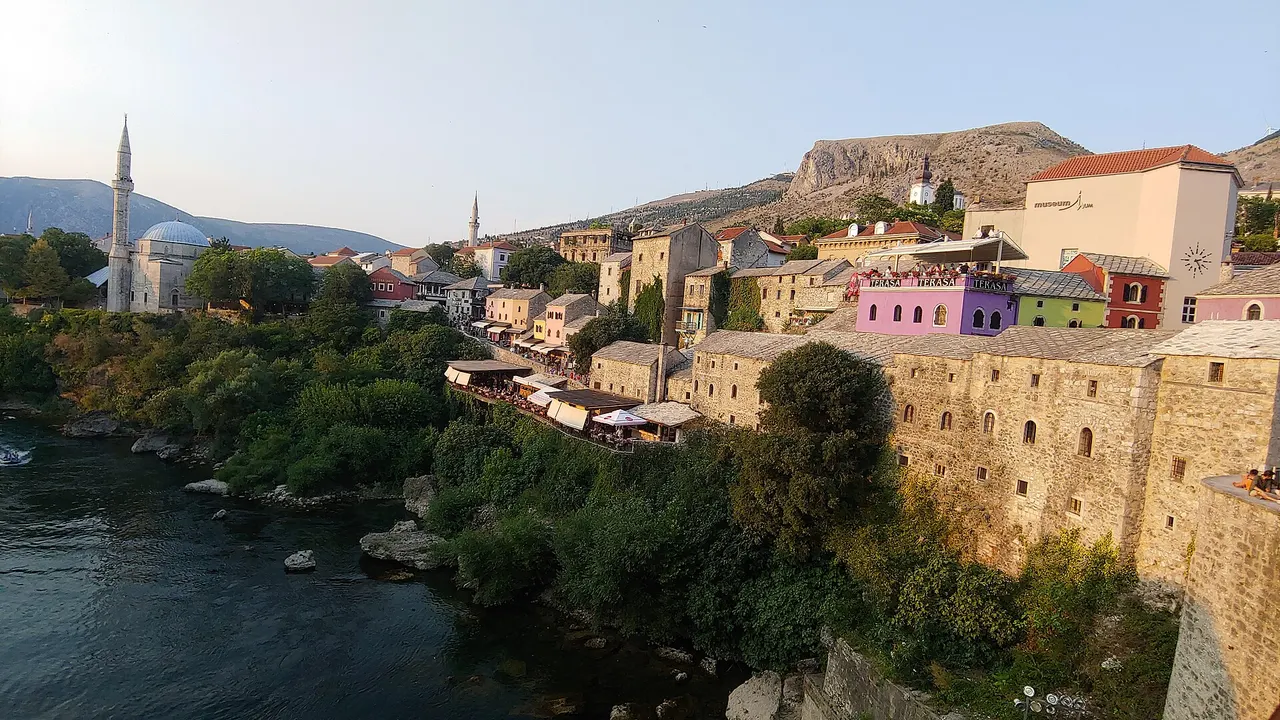 Mostar Old Town at sunset, stone roofs and mosque minarets above the Neretva, with restaurant terraces and TERASA signs along the riverbank