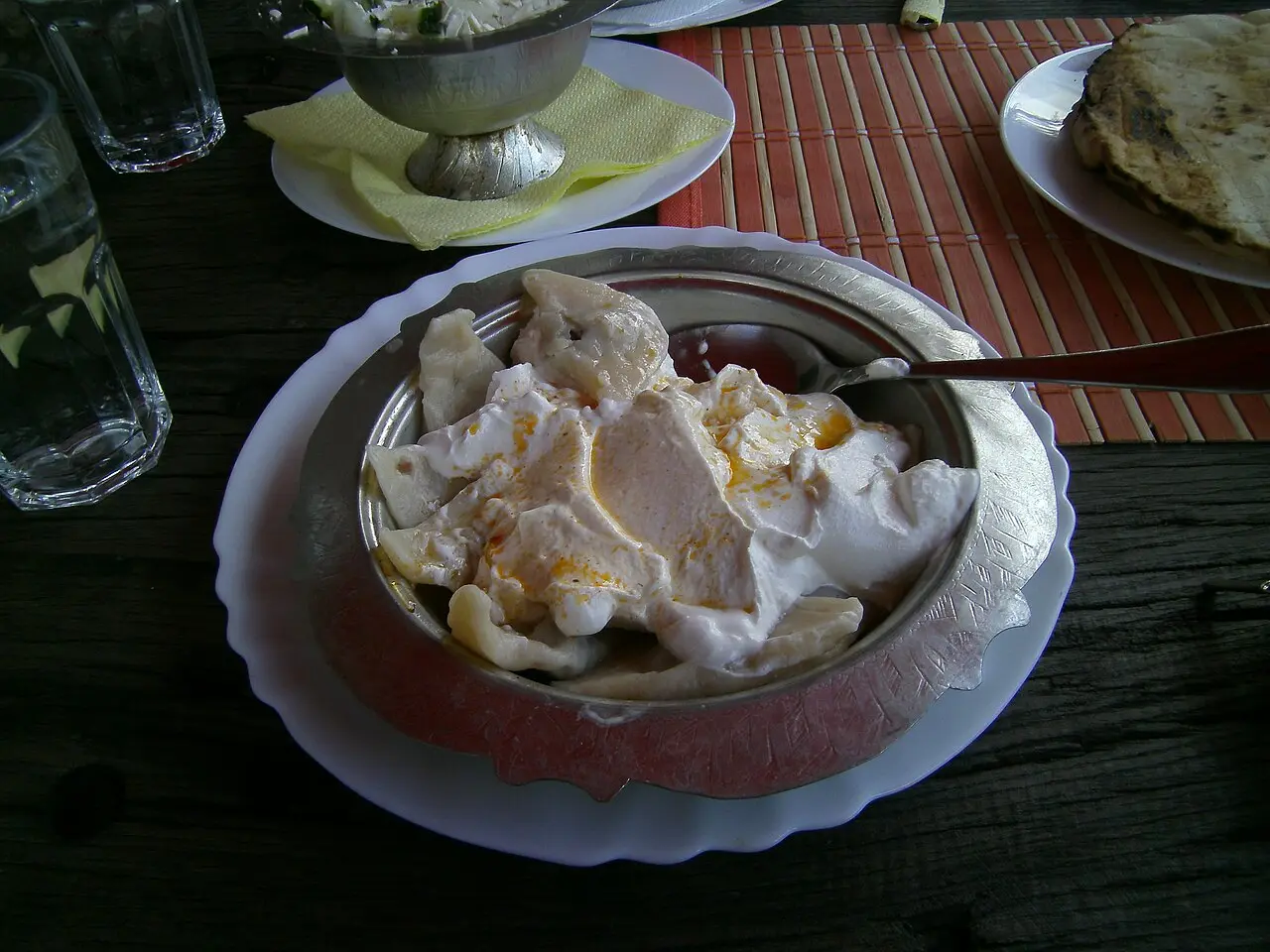 A metal bowl of klepe dumplings buried under garlic yoghurt with a spoon, a plate of flatbread and glasses of water on the table