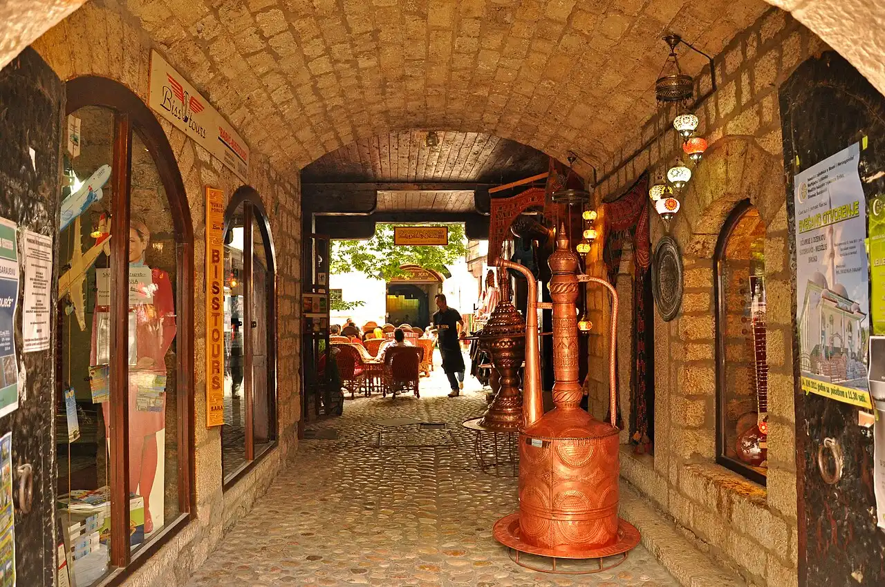 The vaulted stone passage into the courtyard of Morica Han in Sarajevo, hung with lamps and lined with copper stills, tables visible at the far end
