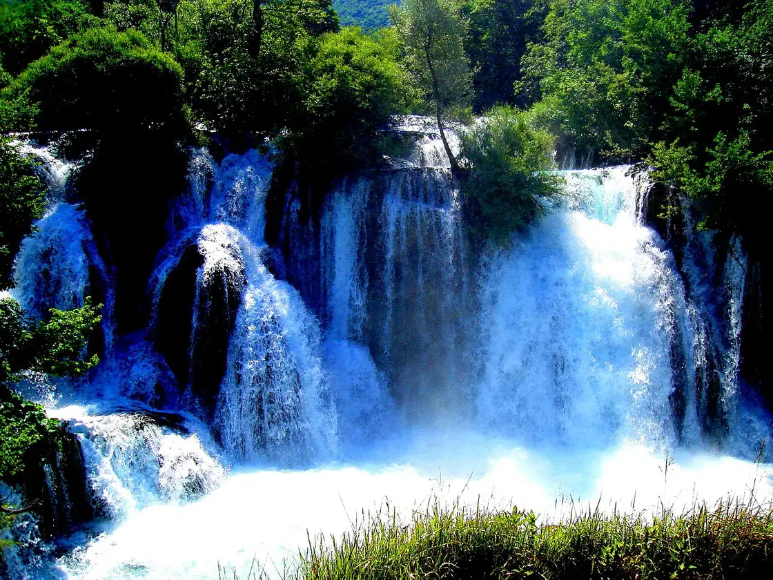 Wide low waterfalls of the Una spilling past houses and greenery at Martin Brod