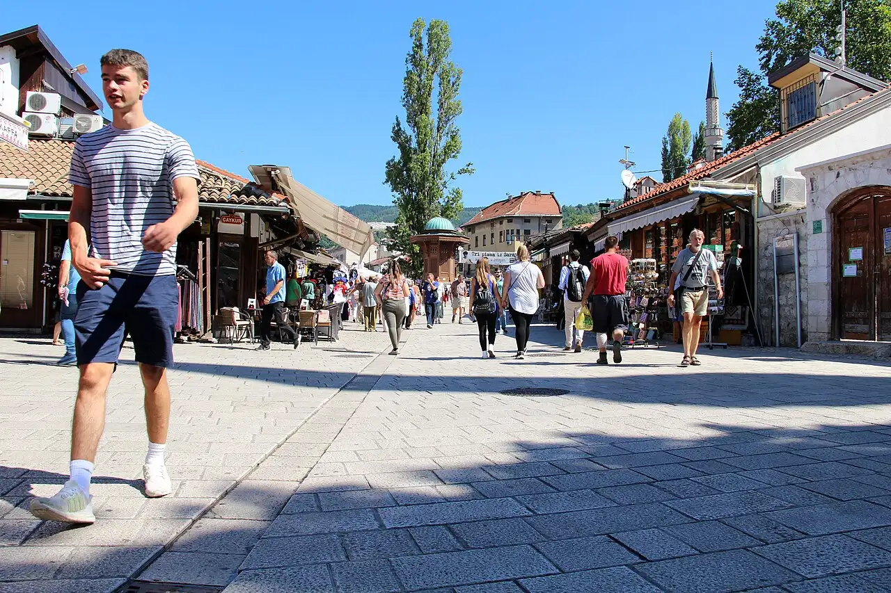 People walking through the pedestrian bazaar of Baščaršija in Sarajevo past shops, the Sebilj fountain and a minaret