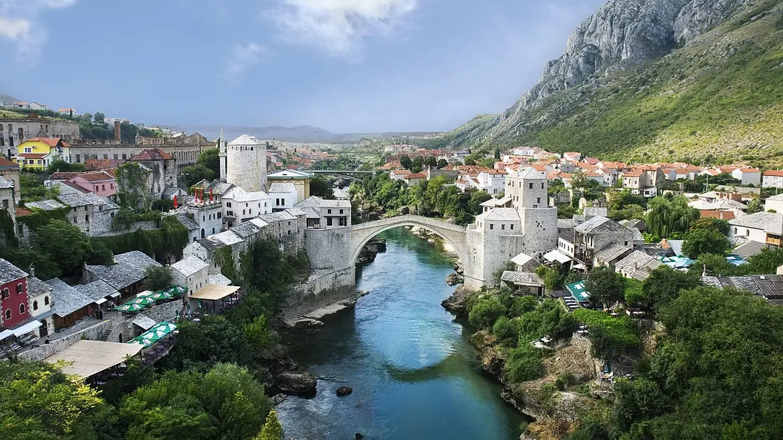 The Old Bridge of Mostar over the green Neretva, with bazaar cafes and stone houses on the east bank and the modern city climbing the west bank