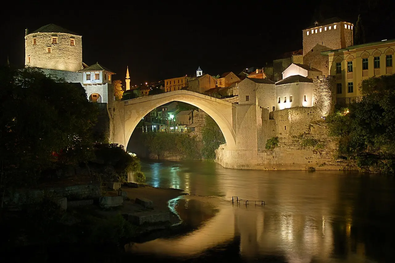 Stari Most and its two stone towers floodlit at night above a quiet, mirror-still Neretva in Mostar, with no crowds